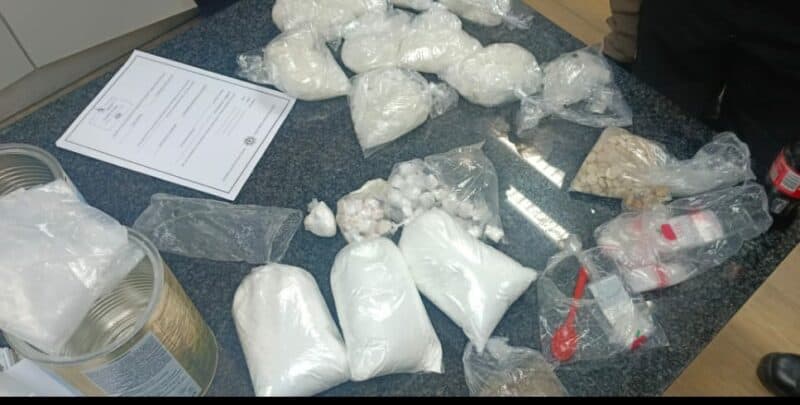 Bags of white powder and plastic-covered bundles are spread across a dark surface, suggesting a police evidence display. A paper with text is visible.