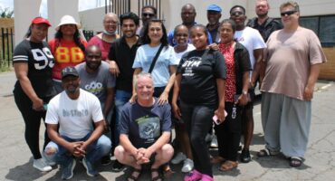 diverse group of 16 people standing and smiling outdoors in front of a light-colored building. They convey a sense of community and friendship.