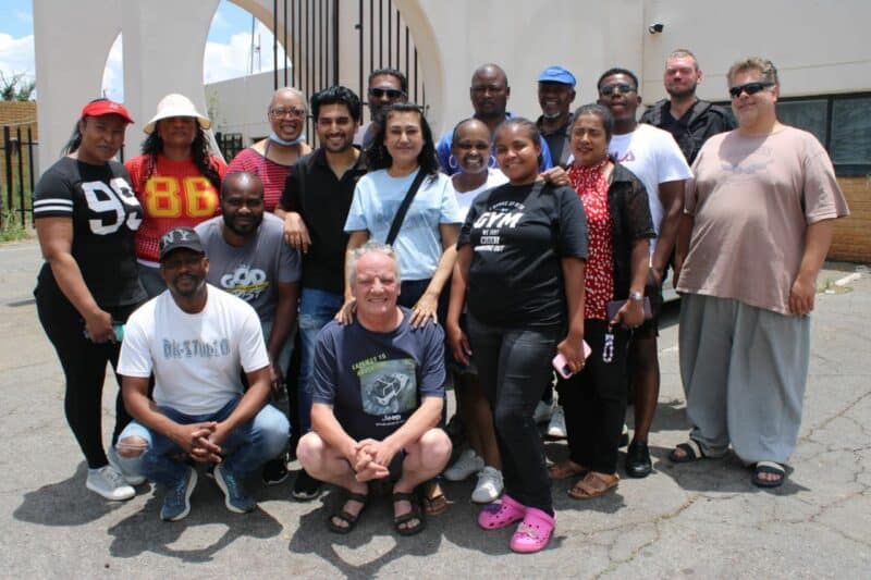diverse group of 16 people standing and smiling outdoors in front of a light-colored building. They convey a sense of community and friendship.