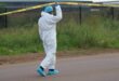 A person in a white protective suit and blue shoe covers lifts yellow crime scene tape on a road. The background features a grassy area and a fence.