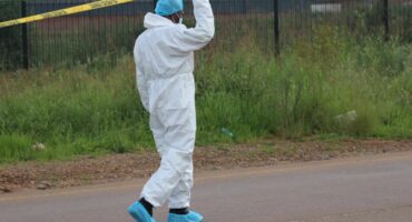 A person in a white protective suit and blue shoe covers lifts yellow crime scene tape on a road. The background features a grassy area and a fence.