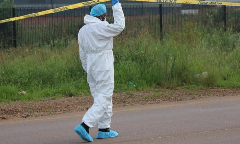 A person in a white protective suit and blue shoe covers lifts yellow crime scene tape on a road. The background features a grassy area and a fence.