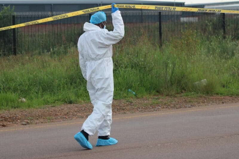 A person in a white protective suit and blue shoe covers lifts yellow crime scene tape on a road. The background features a grassy area and a fence.