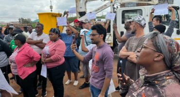 A diverse group of people holds up signs during a protest, with determined expressions. A cloudy sky and utility truck are visible in the background.