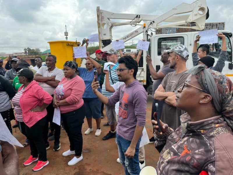 A diverse group of people holds up signs during a protest, with determined expressions. A cloudy sky and utility truck are visible in the background.