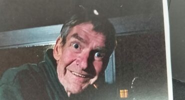 A person with wide eyes and a surprised expression looks through a window at night. Their face is illuminated, creating a mysterious, playful tone.