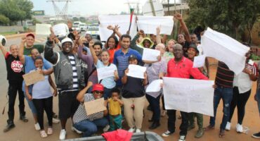 A diverse group of people cheerfully hold protest signs on a roadside, conveying unity and determination. Power lines and trees are visible in the background.