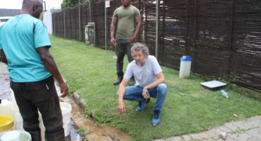 Three men gather outdoors near yellow and white containers on grass, examining something. The mood is serious, suggesting an issue being addressed.