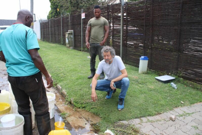 Three men gather outdoors near yellow and white containers on grass, examining something. The mood is serious, suggesting an issue being addressed.
