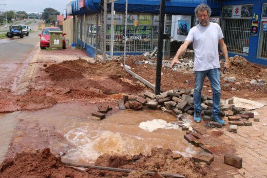 A man in a white shirt and jeans gestures towards a burst water pipe flooding a muddy, brick-strewn street, conveying frustration and urgency.
