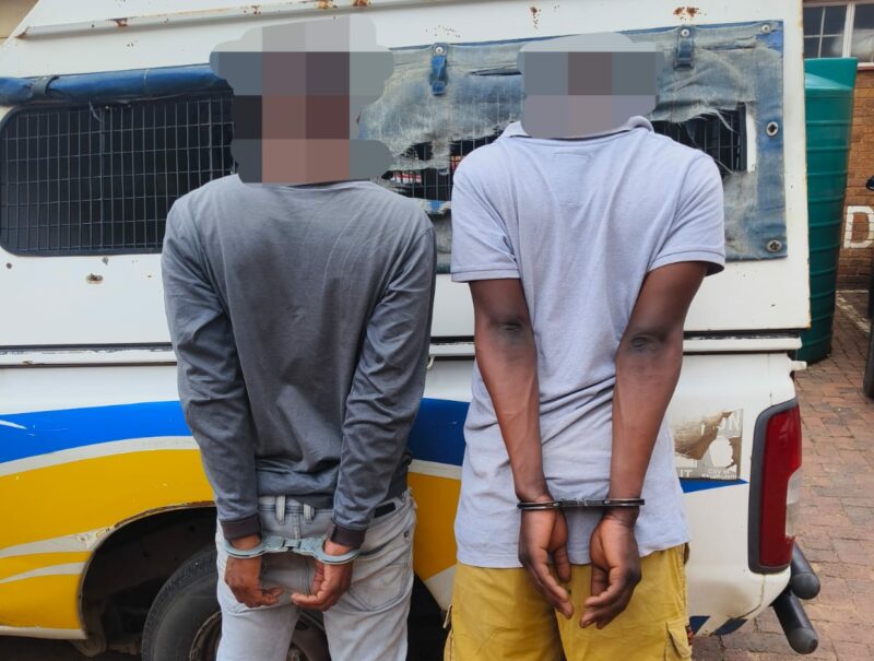 Two individuals in handcuffs stand behind a police vehicle on a brick surface. The scene conveys a serious and somber tone.