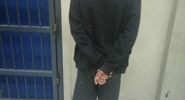 A person in a black hoodie, blurred face, stands with hands clasped behind back. They are in front of a blue metal door, in a tiled room, suggesting a somber atmosphere.