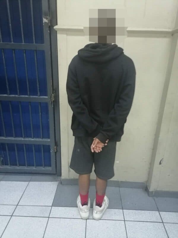 A person in a black hoodie, blurred face, stands with hands clasped behind back. They are in front of a blue metal door, in a tiled room, suggesting a somber atmosphere.