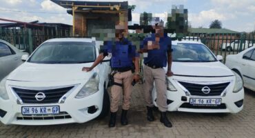 Two individuals in uniform stand beside white Nissan cars in a parking lot under a partly cloudy sky. Their faces are pixelated for privacy.