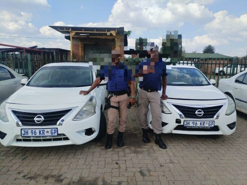 Two individuals in uniform stand beside white Nissan cars in a parking lot under a partly cloudy sky. Their faces are pixelated for privacy.