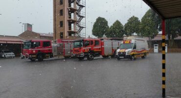 Four emergency vehicles, including fire trucks and an ambulance, are parked in a station courtyard as rain falls heavily, creating a calm, overcast atmosphere.