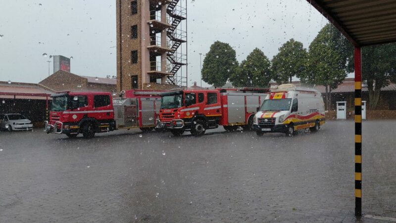 Four emergency vehicles, including fire trucks and an ambulance, are parked in a station courtyard as rain falls heavily, creating a calm, overcast atmosphere.