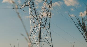 A tall electrical pylon stands against a clear, blue sky, with a few fluffy clouds. Grassy plants frame the foreground, creating a serene scene.