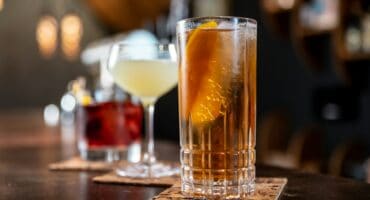 Close-up of three cocktails on a bar counter, each on a coaster. The front drink is a highball with ice and an orange slice, evoking a classy, relaxed vibe.