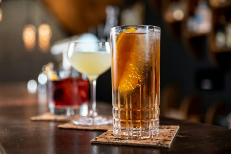 Close-up of three cocktails on a bar counter, each on a coaster. The front drink is a highball with ice and an orange slice, evoking a classy, relaxed vibe.