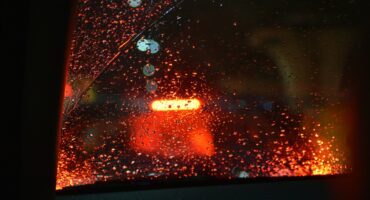 Raindrops on a car window with blurred red taillights in the background, creating a moody, nighttime atmosphere with a sense of calmness.