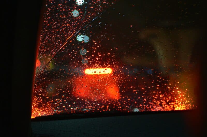 Raindrops on a car window with blurred red taillights in the background, creating a moody, nighttime atmosphere with a sense of calmness.
