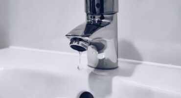 A shiny chrome bathroom faucet with a round handle drips water into a white ceramic sink. The scene feels calm and minimalistic.