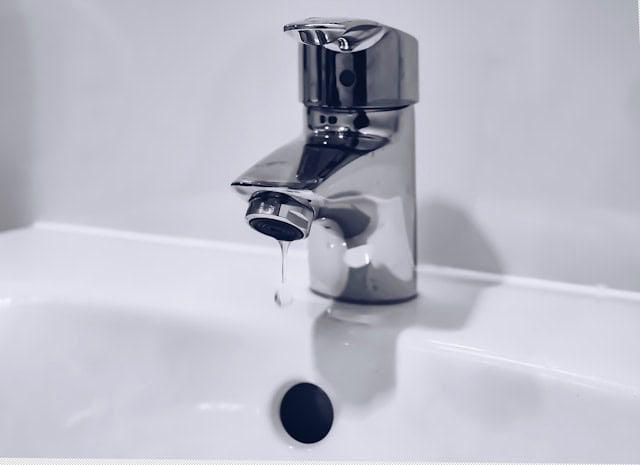 A shiny chrome bathroom faucet with a round handle drips water into a white ceramic sink. The scene feels calm and minimalistic.