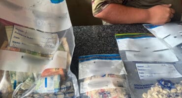 Bags with cash and pills marked "South African Police Service" rest on a dark table. A person in camouflage sits nearby, suggesting a police operation.