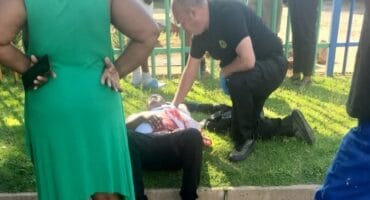A person lies on the grass, receiving medical attention from a responder, surrounded by concerned onlookers near a colorful fence, conveying urgency and concern.