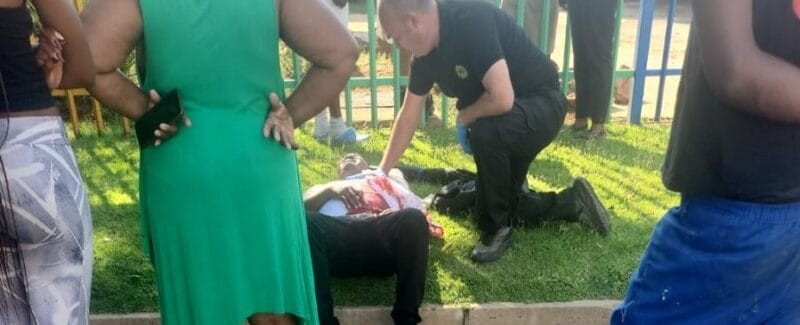 A person lies on the grass, receiving medical attention from a responder, surrounded by concerned onlookers near a colorful fence, conveying urgency and concern.