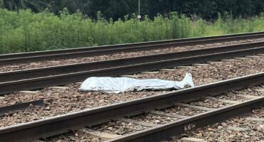 A metallic sheet covers an object on railway tracks surrounded by scattered rocks. Lush green trees and a cloudy sky form the backdrop, invoking tension.