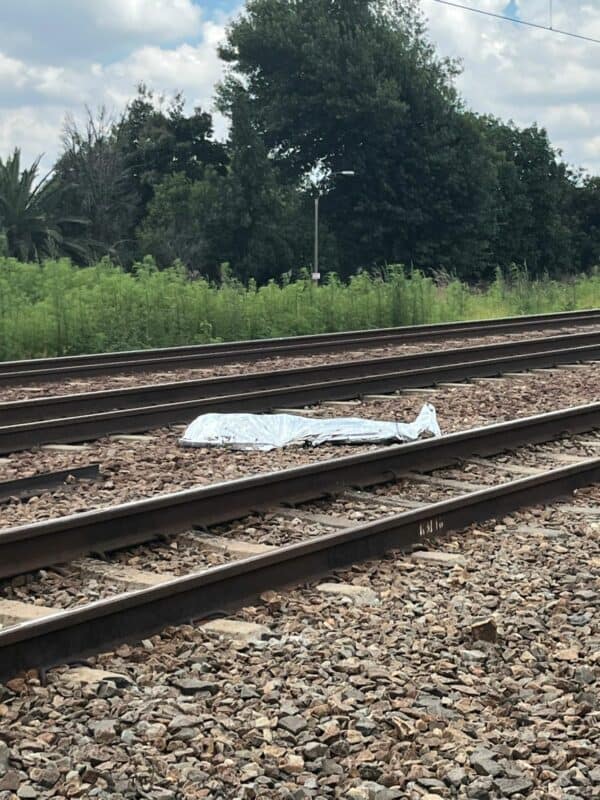 A metallic sheet covers an object on railway tracks surrounded by scattered rocks. Lush green trees and a cloudy sky form the backdrop, invoking tension.