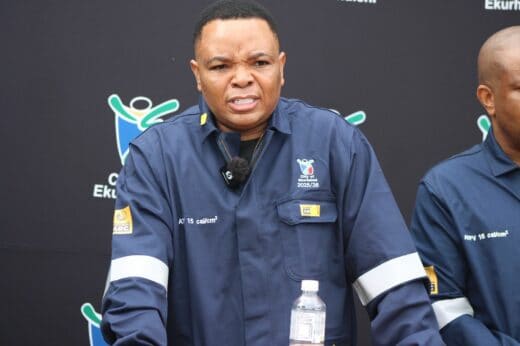 A person in a dark blue work uniform speaks passionately at an event. The background features a logo and text. They hold a water bottle, conveying urgency.