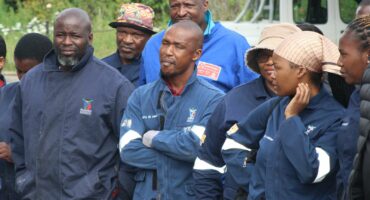 A diverse group of people wearing blue uniforms stands outdoors, some smiling and others reflective. Overcast weather, greenery, and a parked vehicle create a calm atmosphere.
