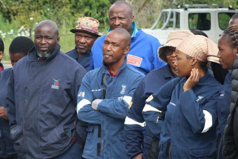 A diverse group of people wearing blue uniforms stands outdoors, some smiling and others reflective. Overcast weather, greenery, and a parked vehicle create a calm atmosphere.