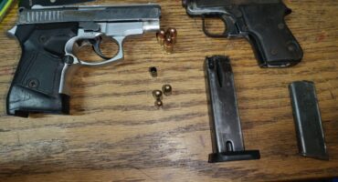 Two pistols and magazines with bullets are placed on a wooden table. Nearby, there are bank cards, hinting at personal belongings. The tone is neutral yet tense.