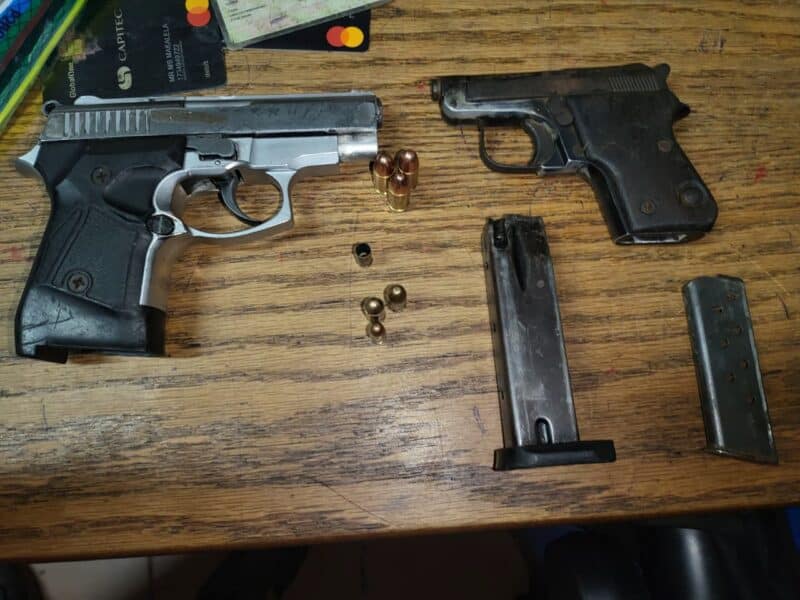 Two pistols and magazines with bullets are placed on a wooden table. Nearby, there are bank cards, hinting at personal belongings. The tone is neutral yet tense.