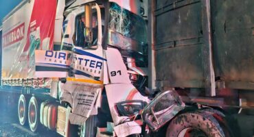 A nighttime scene depicting a collision between two large trucks on a wet road. The front of a white truck is heavily damaged, conveying a sense of impact.
