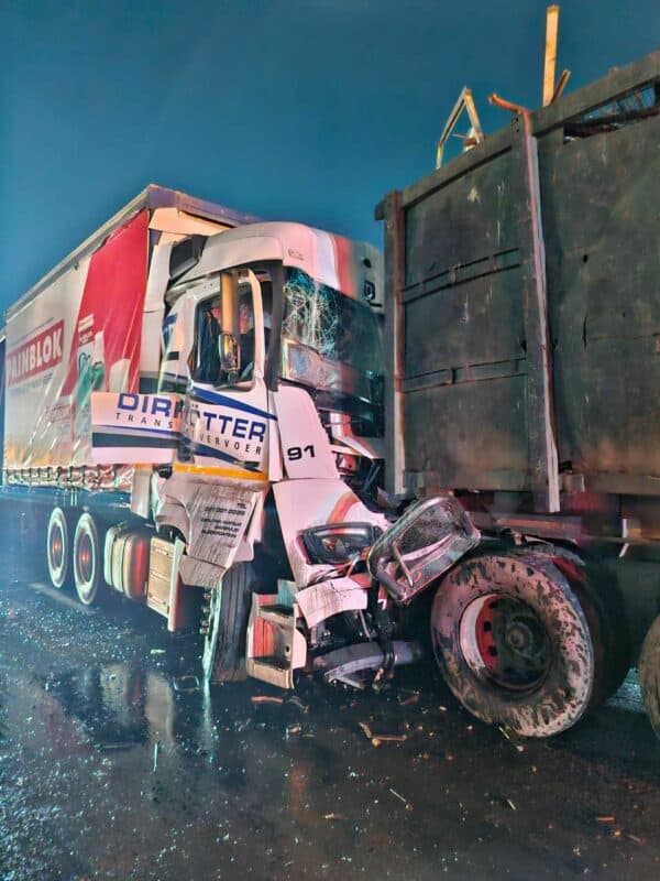 A nighttime scene depicting a collision between two large trucks on a wet road. The front of a white truck is heavily damaged, conveying a sense of impact.