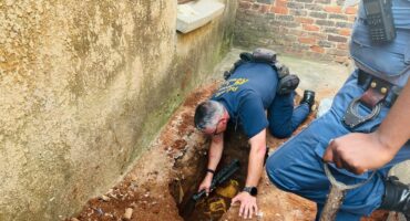 A police officer crouches, inspecting a dug-up hole near a building's wall, holding a tool. Another officer stands by with a shovel. The scene conveys investigation and discovery.