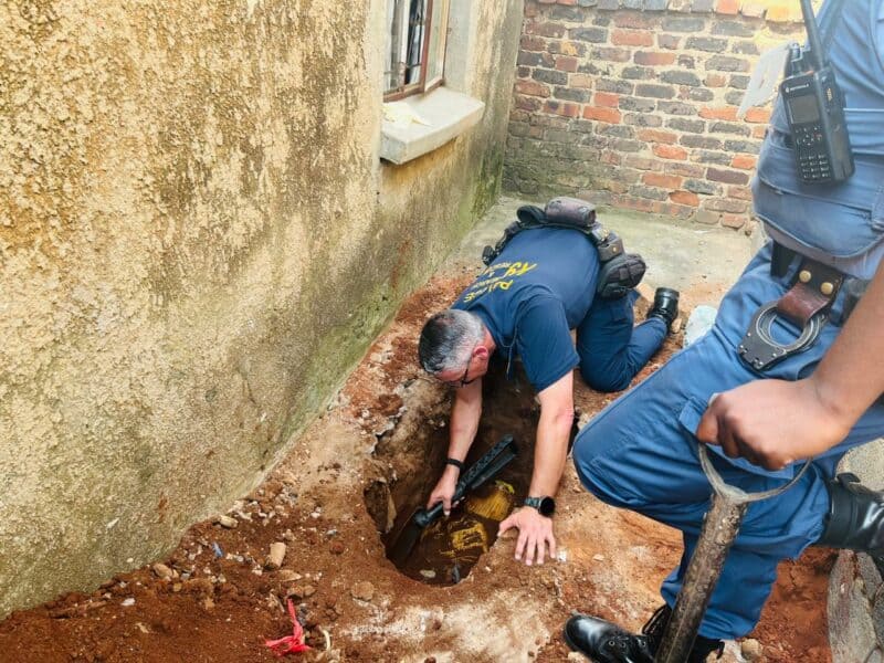 A police officer crouches, inspecting a dug-up hole near a building's wall, holding a tool. Another officer stands by with a shovel. The scene conveys investigation and discovery.