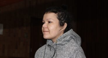 A person with a calm expression wearing a gray hoodie stands in a dimly lit indoor setting, possibly a courtroom. The tone is serious and contemplative.