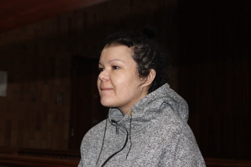 A person with a calm expression wearing a gray hoodie stands in a dimly lit indoor setting, possibly a courtroom. The tone is serious and contemplative.
