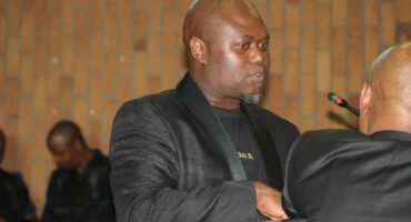 A man in a black suit speaks seriously into a microphone at an indoor event. Others sit in the background against a brick wall, creating a formal atmosphere.