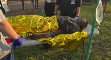 Emergency responders in masks tend to an injured person on a stretcher covered in a gold thermal blanket, outdoors on grass near a road.