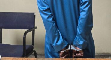 A person in a blue shirt, handcuffed, stands facing away next to a chair. A wooden table displays small bags of an unknown substance. The scene suggests a serious and tense mood.