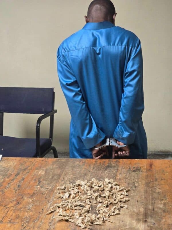 A person in a blue shirt, handcuffed, stands facing away next to a chair. A wooden table displays small bags of an unknown substance. The scene suggests a serious and tense mood.