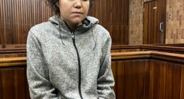 A person sits in a wooden courtroom, appearing concerned and pensive. They wear a grey hoodie, with wood paneling visible in the background.
