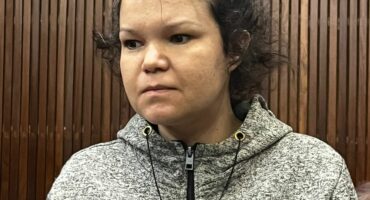 A person with a neutral expression stands in a room with wooden paneling, wearing a gray zip-up hoodie. The mood appears calm and contemplative.